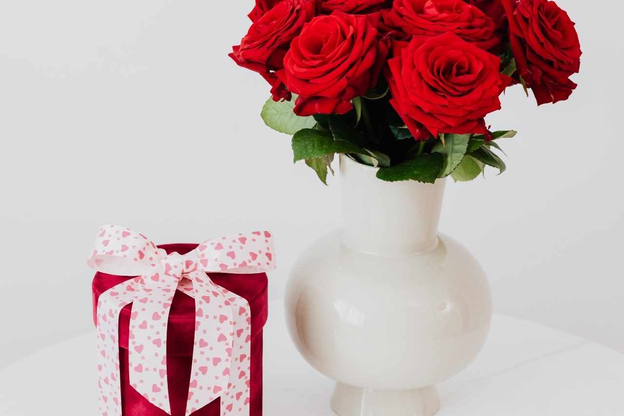 A bouquet of red roses in a white vase on a white table next to a red velvet cylinder box with a white bow with pink hearts on it