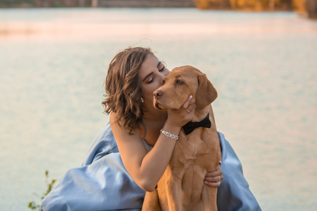 A woman wearing a long blue gown and a detailed diamond bracelet kissing the side of a dog’s head
