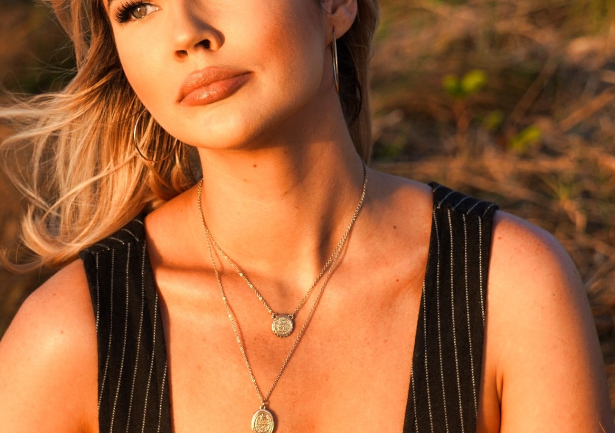 A woman sitting in the sun, wearing two layered gold necklaces with coin pendants