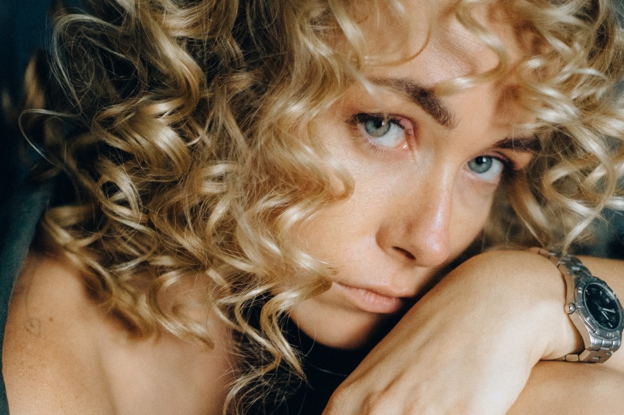 A blonde woman with curly hair poses with a stainless steel watch on her wrist.