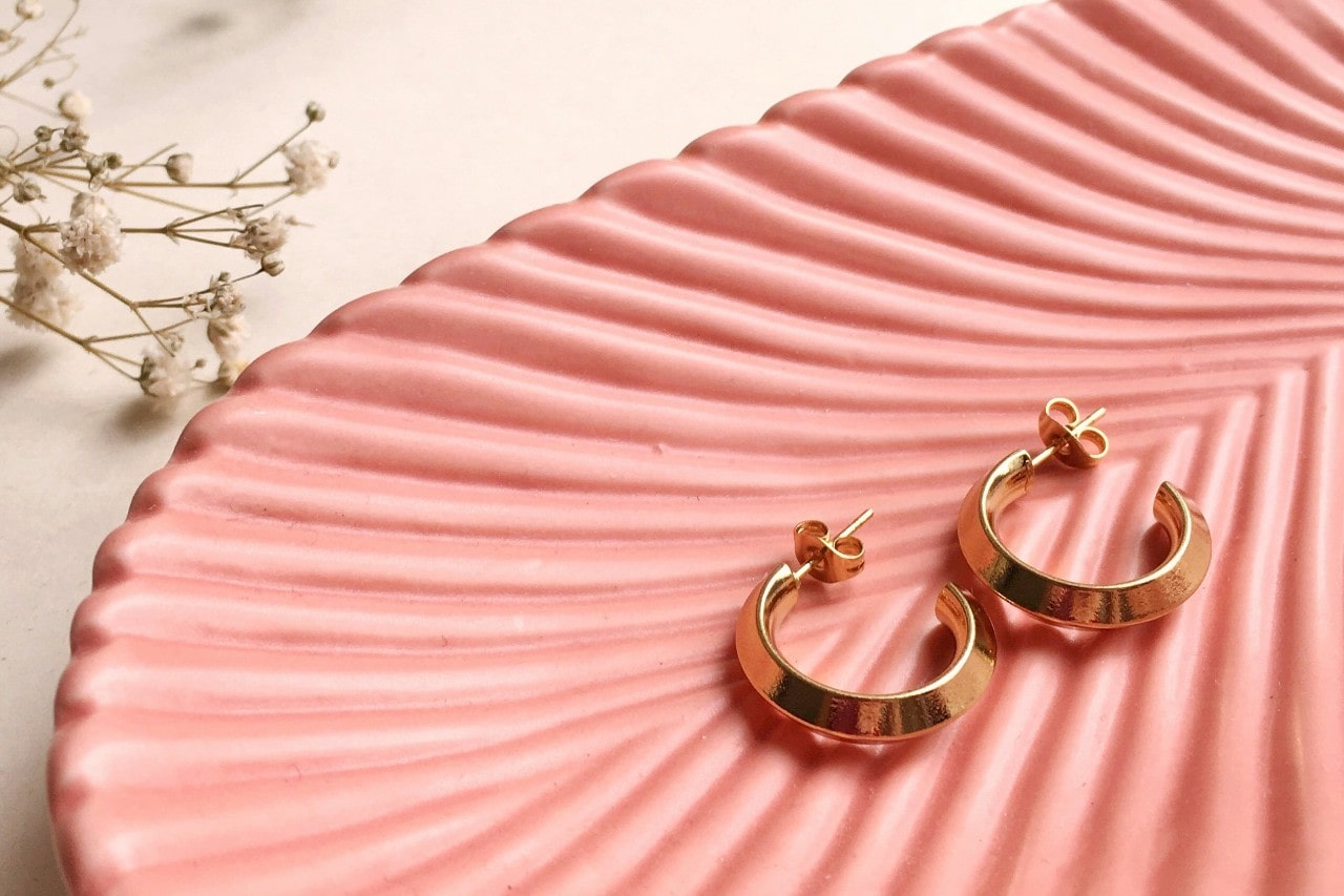 a pair of yellow gold huggie earrings on a pink dish.