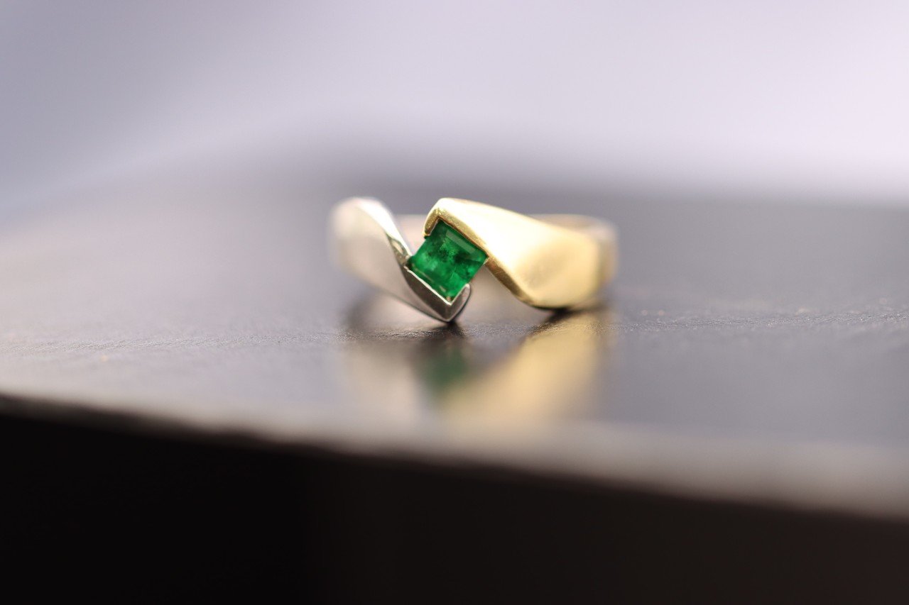 A yellow gold ring featuring a princess cut emerald on a metallic surface.