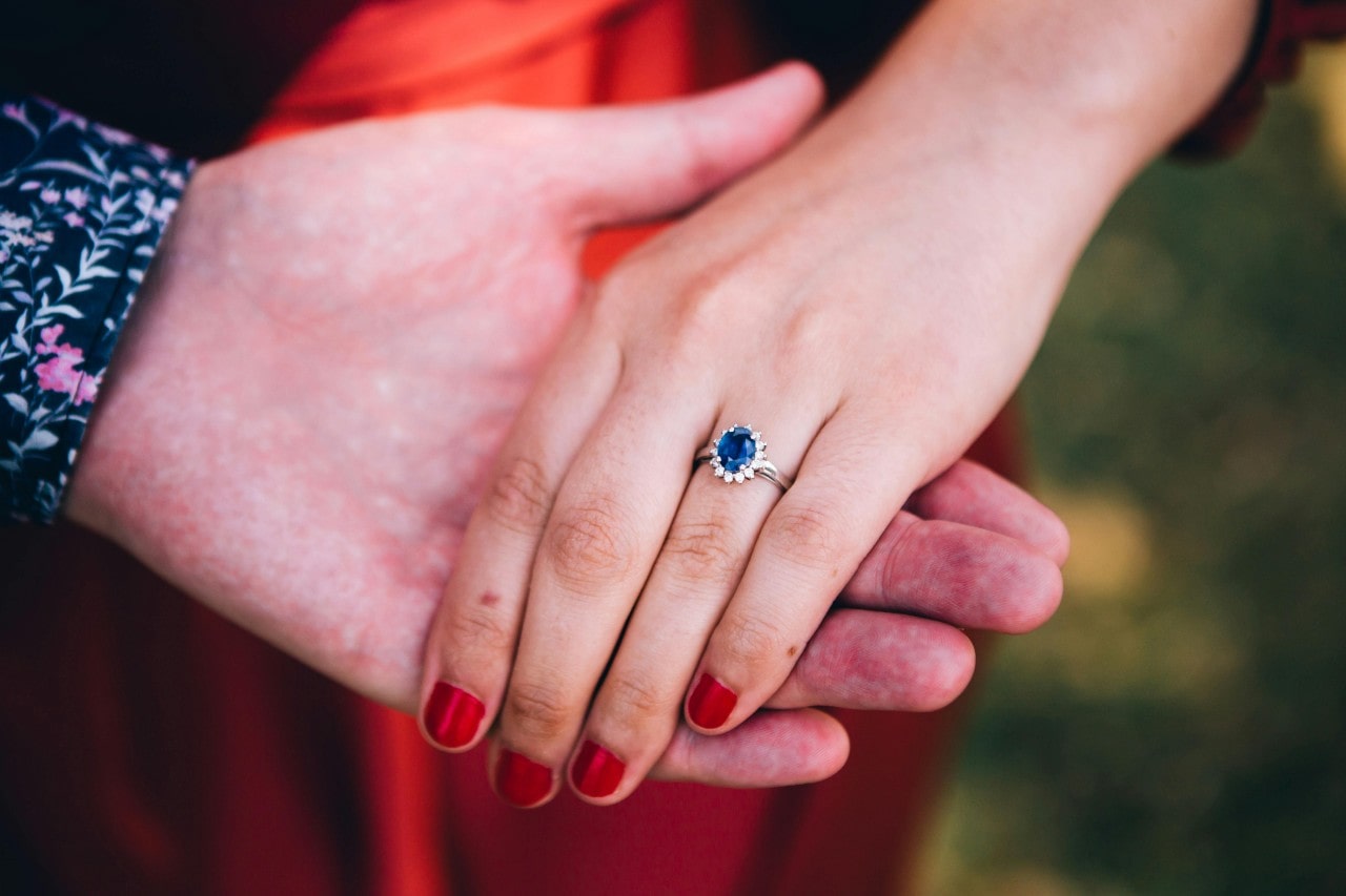 A man’s hand holding a woman’s hand, adorned with a sapphire engagement ring.