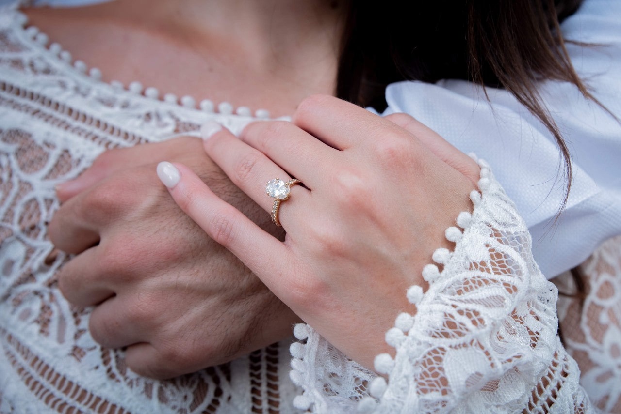 A close-up of a woman showcasing her oval diamond engagement ring as she holds man’s hand to her heart, while wearing white blouse. A close-up of a woman showcasing her oval diamond engagement ring as she holds man’s hand to her heart, while wearing white blouse.
