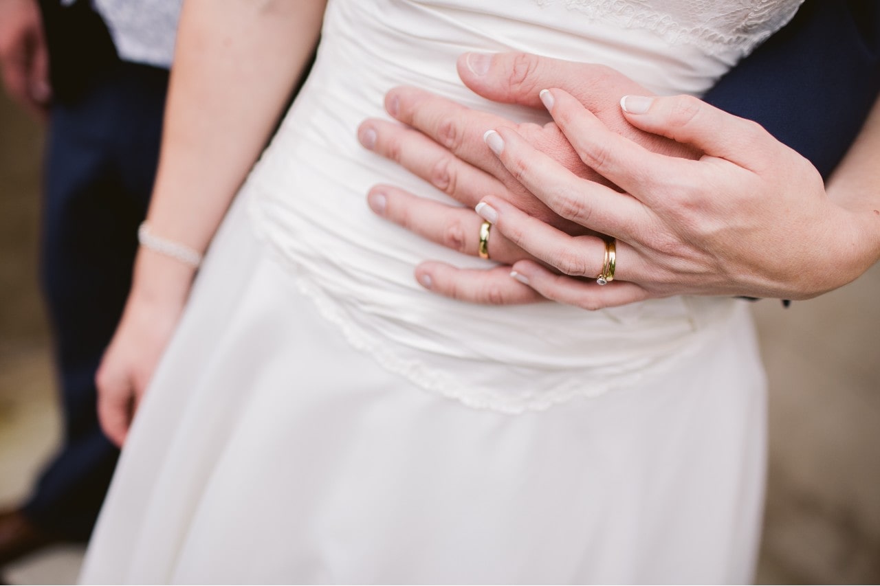 A close-up of a groom embracing his bride from behind, both wearing gold wedding bands.