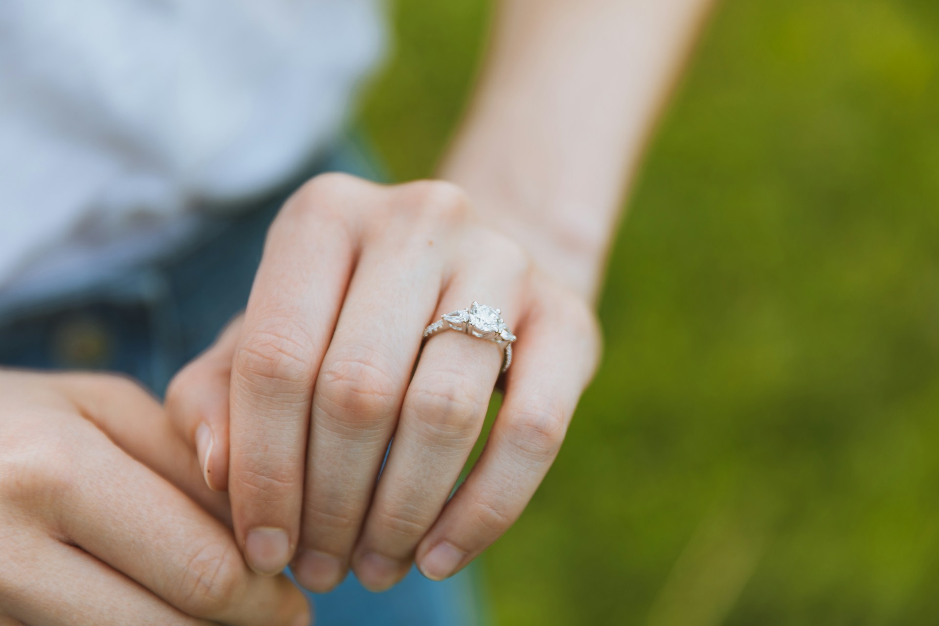 A white gold three stone engagement ring displayed on a woman’s hand against a blurry grass A white gold three stone engagement ring displayed on a woman’s hand against a blurry grass