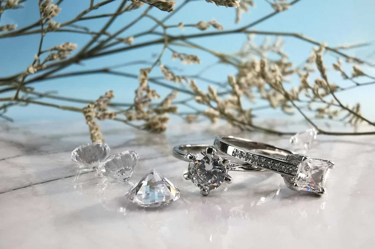 A close-up of two stunning diamond rings next to 3 round cut diamonds on a beachy backdrop.